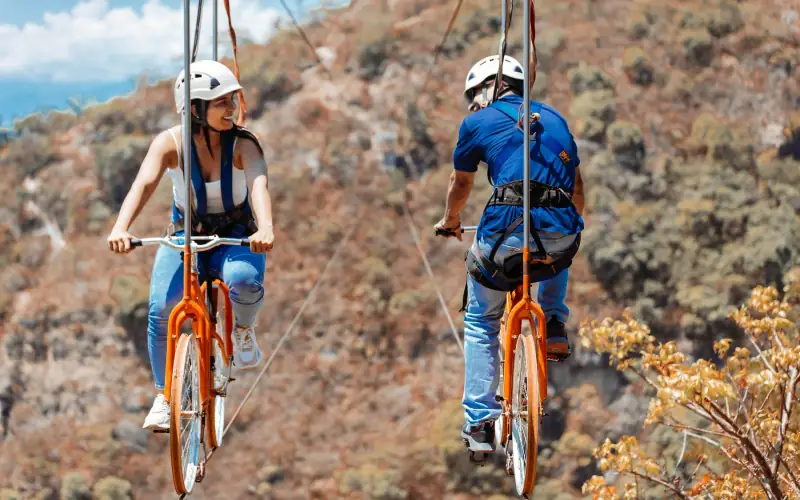 Dos personas con casco pedaleando bicicletas colgadas de un cable sobre la montaña